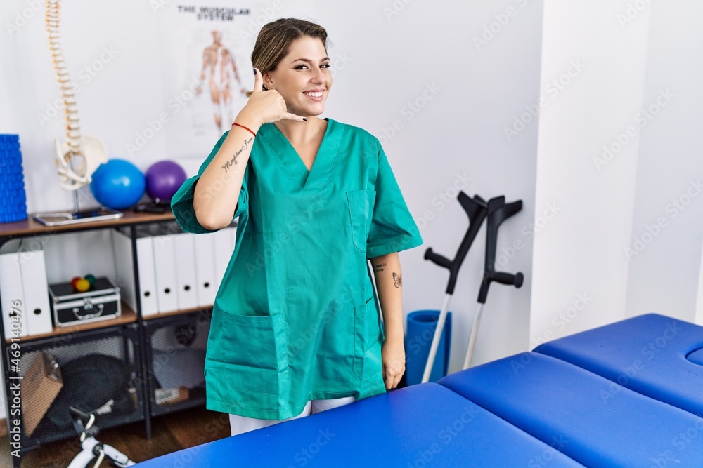 Young hispanic woman wearing physiotherapist uniform standing at clinic smiling doing phone gesture with hand and fingers like talking on the telephone. communicating concepts. Young hispanic woman wearing physiotherapist uniform standing at clinic smiling doing phone gesture with hand and fingers like talking on the telephone. communicating concepts.