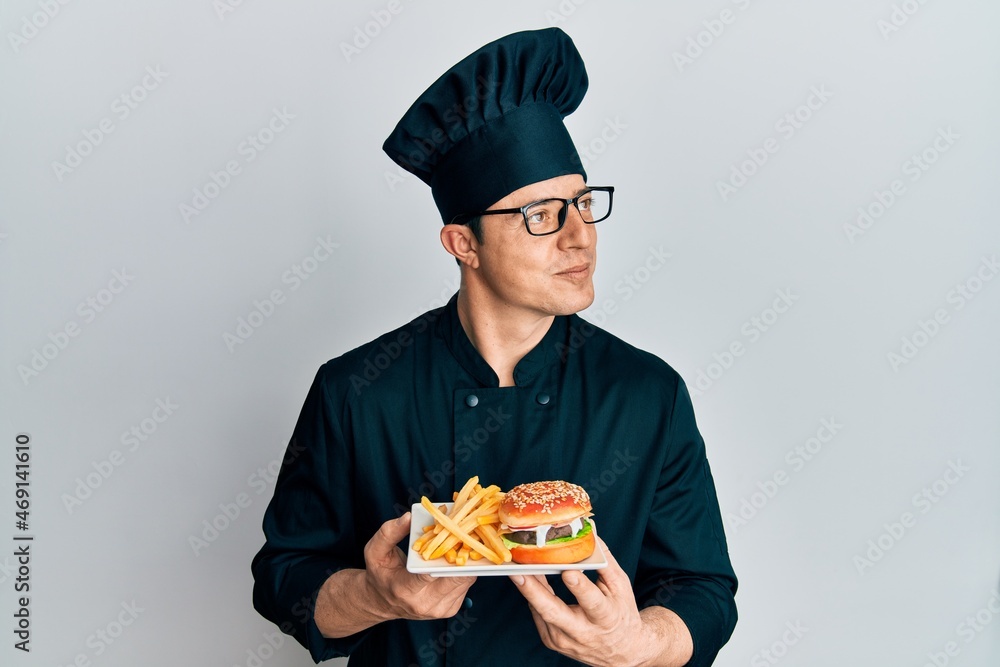 Handsome young man chef holding burger with fries smiling looking to the side and staring away thinking. Handsome young man chef holding burger with fries smiling looking to the side and staring away thinking.
