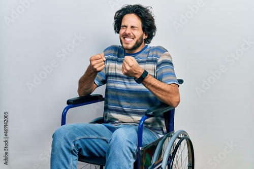 Handsome hispanic man sitting on wheelchair excited for success with arms raised and eyes closed celebrating victory smiling. winner concept.