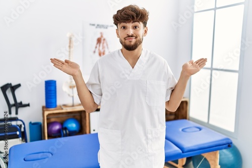 Young arab man working at pain recovery clinic clueless and confused expression with arms and hands raised. doubt concept.