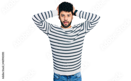 Young arab man with beard wearing casual striped sweater crazy and scared with hands on head, afraid and surprised of shock with open mouth