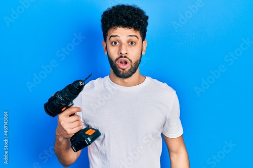 Young arab man with beard using drill scared and amazed with open mouth for surprise, disbelief face