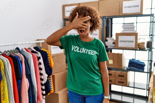 Young african american woman wearing volunteer t shirt at donations stand peeking in shock covering face and eyes with hand, looking through fingers with embarrassed expression.