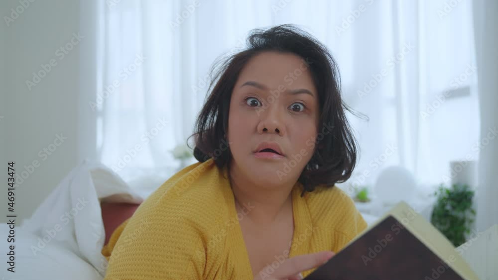 Asian woman oversize big chubby girl lying in bed reading a book in the bedroom showing a panic look at the camera. Leisure and hobbies on holiday. Asian woman oversize big chubby girl lying in bed reading a book in the bedroom showing a panic look at the camera. Leisure and hobbies on holiday.