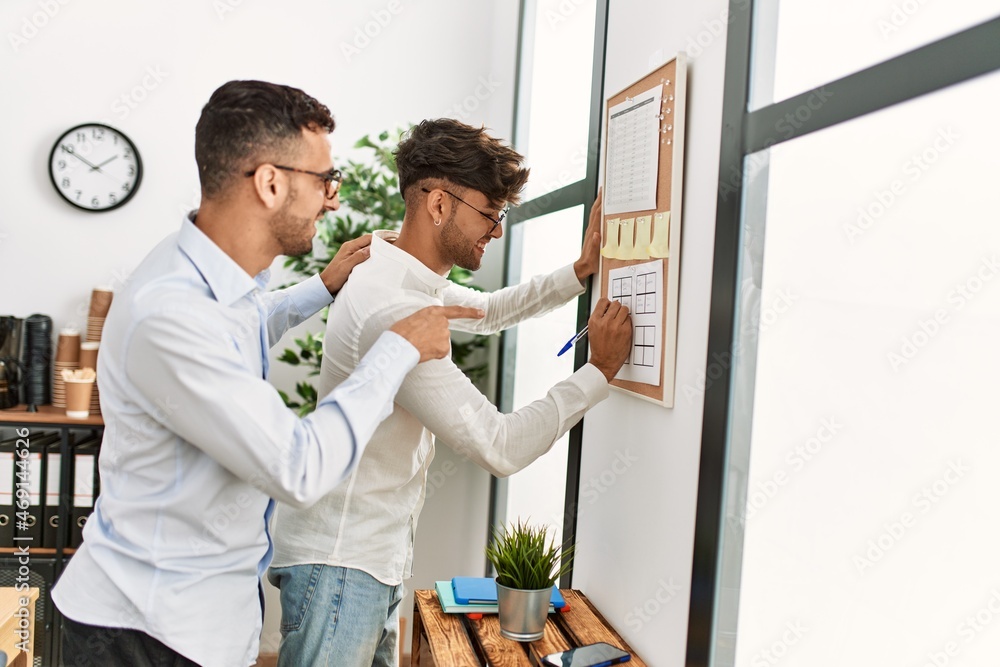 Two hispanic men couple writing on corkboard working at office Two hispanic men couple writing on corkboard working at office