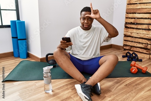 Young african man sitting on training mat at the gym using smartphone making fun of people with fingers on forehead doing loser gesture mocking and insulting.