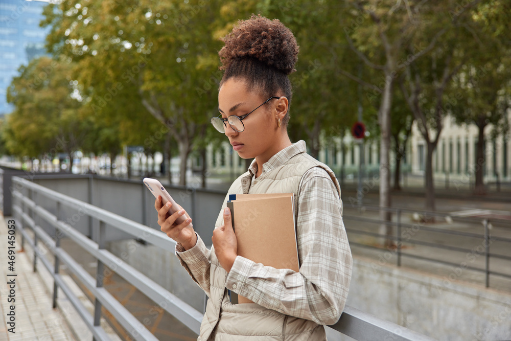 Fototapeta premium Intelligent female student concentrated at smartphone screen learns schedule for next week holds textbook wears round spectacles and casual clothes poses at city against trees uses modern technologies