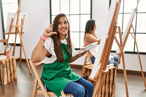 Young hispanic artist women painting on canvas at art studio smiling doing phone gesture with hand and fingers like talking on the telephone. communicating concepts.