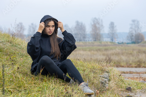 A beautiful girl with long hair in black clothes sits on the grass on the outskirts of the city. She puts a hood over her head and looks away
