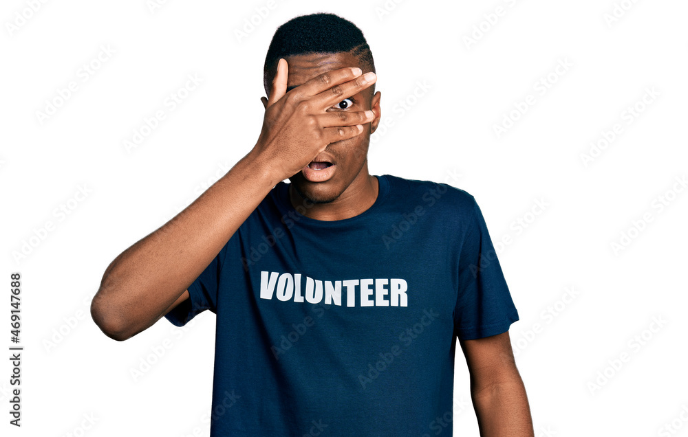 Fototapeta premium Young african american man wearing volunteer t shirt peeking in shock covering face and eyes with hand, looking through fingers with embarrassed expression.