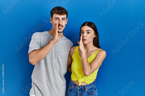 Young hispanic couple standing together over blue background hand on mouth telling secret rumor, whispering malicious talk conversation