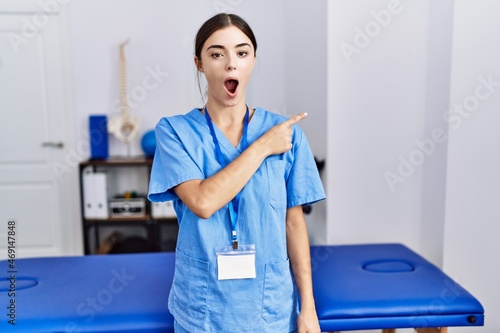 Фотография Young hispanic woman wearing physiotherapist uniform standing at clinic surprised pointing with finger to the side, open mouth amazed expression