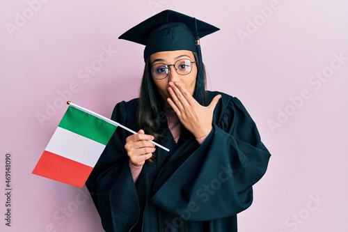 Young hispanic woman wearing graduation uniform holding italy flag covering mouth with hand, shocked and afraid for mistake. surprised expression