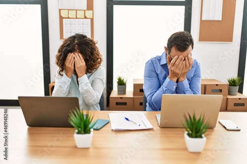 Middle age hispanic woman and man sitting with laptop at the office with sad expression covering face with hands while crying. depression concept.