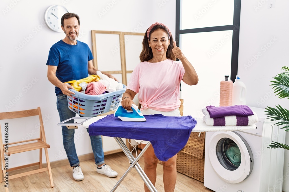 Middle age couple ironing clothes at home smiling happy and positive, thumb up doing excellent and approval sign Middle age couple ironing clothes at home smiling happy and positive, thumb up doing excellent and approval sign