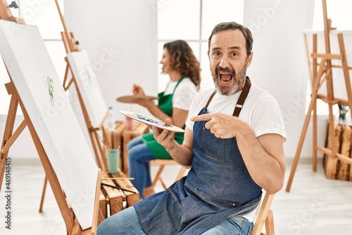 Hispanic middle age man and mature woman at art studio smiling happy pointing with hand and finger