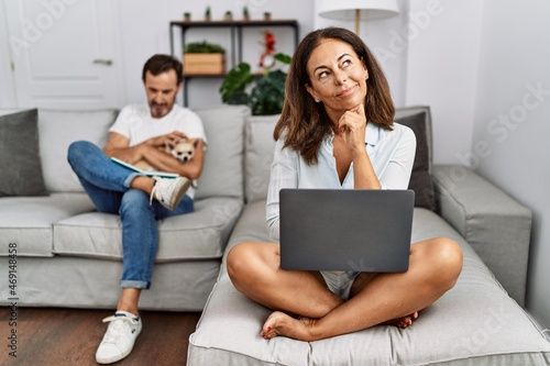 Hispanic middle age couple at home, woman using laptop with hand on chin thinking about question, pensive expression. smiling with thoughtful face. doubt concept.