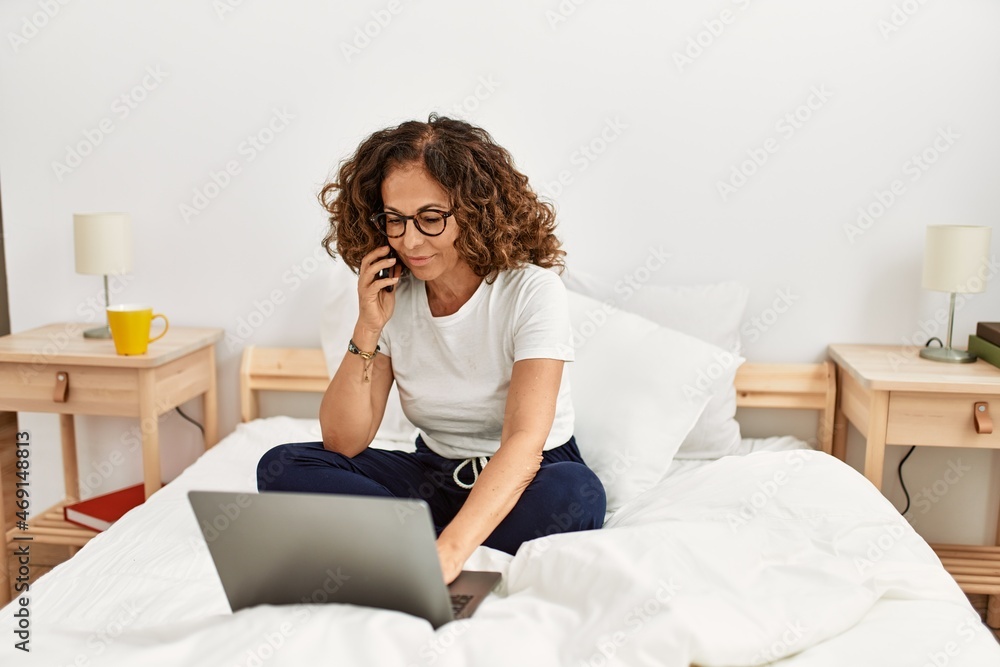 Middle age hispanic woman using laptop talking on the smartphone at bedroom Middle age hispanic woman using laptop talking on the smartphone at bedroom
