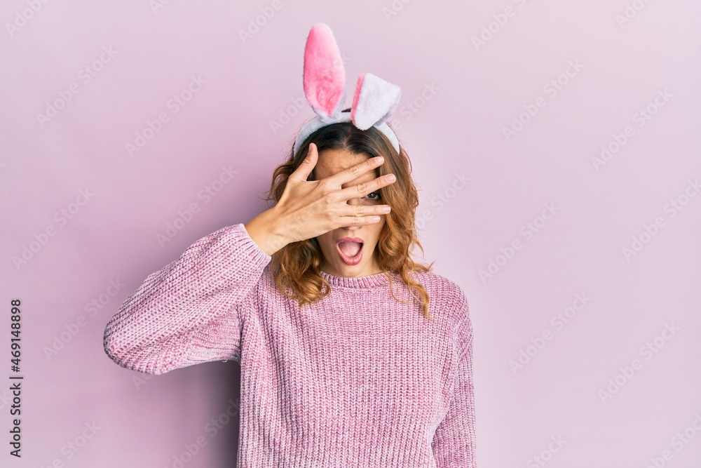Young caucasian woman wearing cute easter bunny ears peeking in shock covering face and eyes with hand, looking through fingers afraid