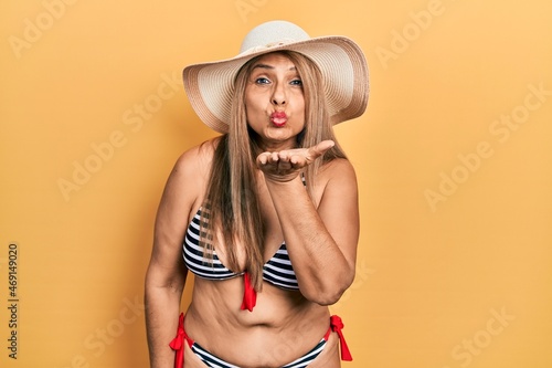 Middle age hispanic woman wearing bikini and summer hat looking at the camera blowing a kiss with hand on air being lovely and sexy. love expression.