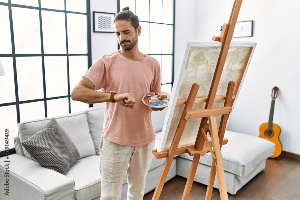 Young hispanic man with beard painting on canvas at home checking the time on wrist watch, relaxed and confident