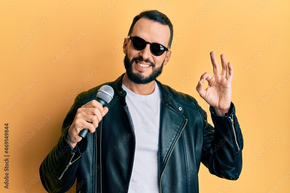 Obraz premium Young man with beard singing song using microphone doing ok sign with fingers, smiling friendly gesturing excellent symbol
