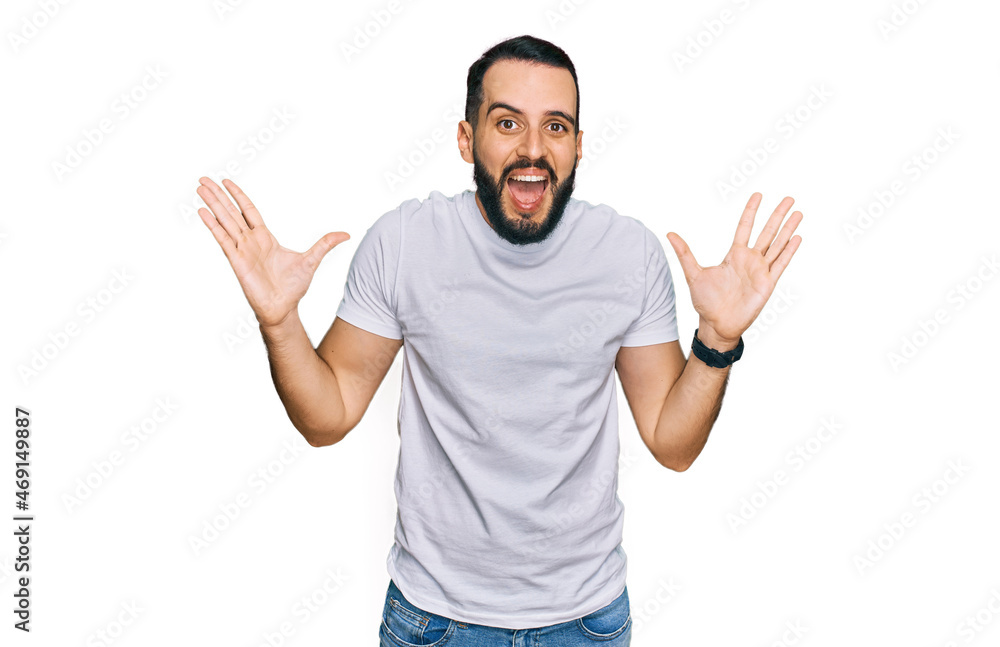 Young man with beard wearing casual white t shirt celebrating crazy and amazed for success with arms raised and open eyes screaming excited. winner concept Young man with beard wearing casual white t shirt celebrating crazy and amazed for success with arms raised and open eyes screaming excited. winner concept