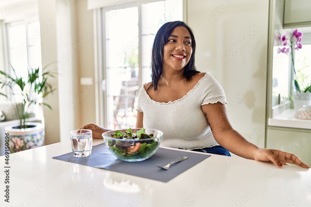 Young hispanic woman eating healthy salad at home looking away to side with smile on face, natural expression. laughing confident. Young hispanic woman eating healthy salad at home looking away to side with smile on face, natural expression. laughing confident.