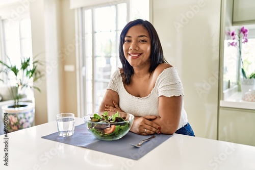 Young hispanic woman eating healthy salad at home happy face smiling with crossed arms looking at the camera. positive person.