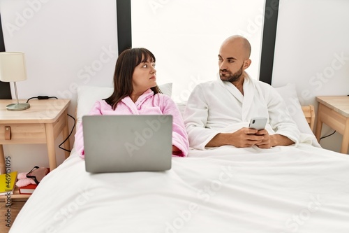 Young hispanic couple using laptop and smartphone sitting on the bed at bedroom.