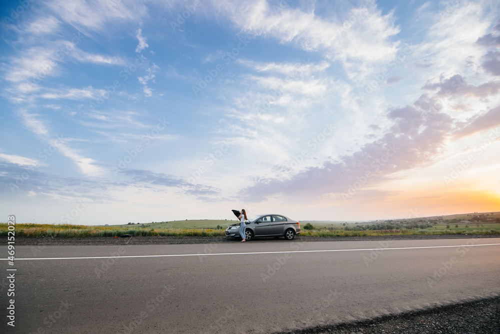 A young girl stands near a broken car in the middle of the highway during sunset and tries to repair it. Breakdown and repair of the car. Troubleshooting the problem. A young girl stands near a broken car in the middle of the highway during sunset and tries to repair it. Breakdown and repair of the car. Troubleshooting the problem.