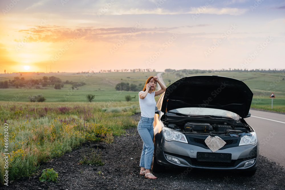 A young girl stands near a broken-down car in the middle of the highway during sunset and tries to call for help on the phone. Breakdown and repair of the car. Waiting for help.