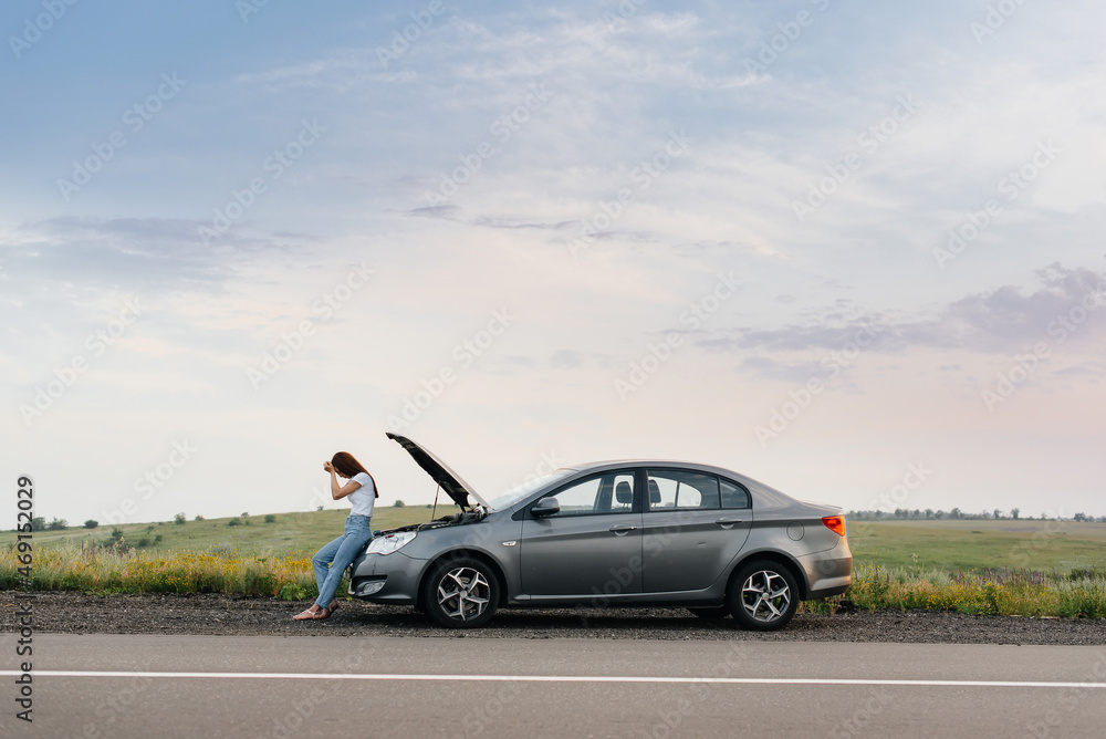 A frustrated young girl stands near a broken-down car in the middle of the highway during sunset. Breakdown and repair of the car. Waiting for help.