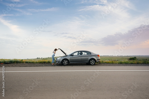 A young girl stands near a broken-down car in the middle of the highway during sunset and tries to call for help on the phone. Breakdown and repair of the car. Waiting for help. © Andrii