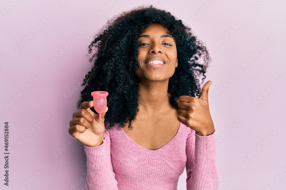 African american woman with afro hair holding menstrual cup smiling happy and positive, thumb up doing excellent and approval sign African american woman with afro hair holding menstrual cup smiling happy and positive, thumb up doing excellent and approval sign