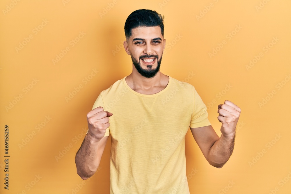 Handsome man with beard wearing casual yellow t shirt celebrating surprised and amazed for success with arms raised and open eyes. winner concept. Handsome man with beard wearing casual yellow t shirt celebrating surprised and amazed for success with arms raised and open eyes. winner concept.