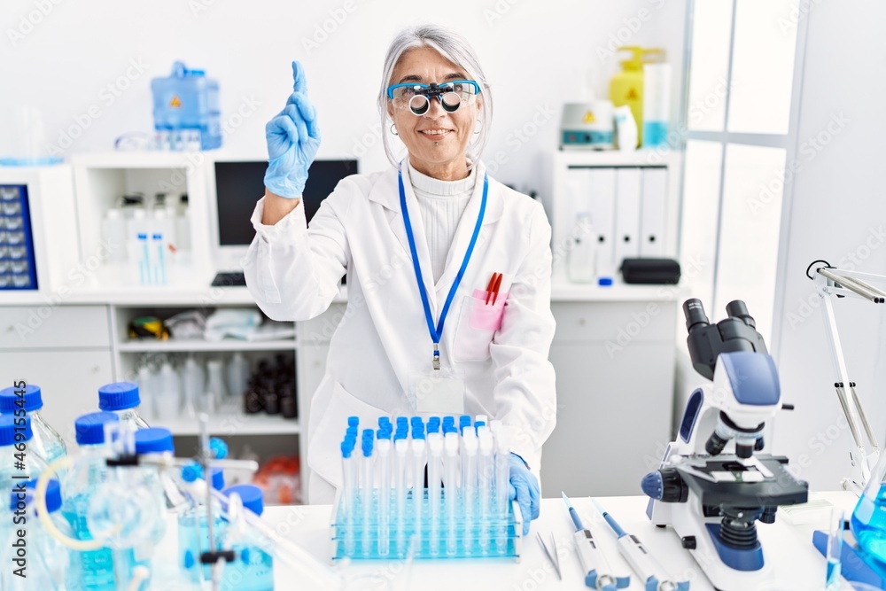Middle age grey-haired woman wearing scientist uniform smiling with an idea or question pointing finger with happy face, number one Middle age grey-haired woman wearing scientist uniform smiling with an idea or question pointing finger with happy face, number one