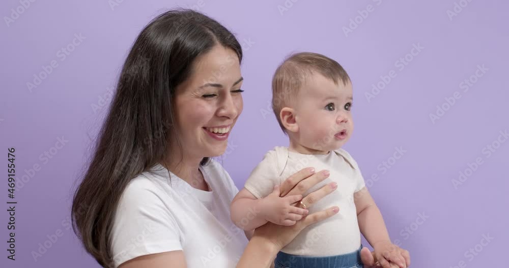 Attractive Caucasian Brunette Female Holding Little Child Girl Daughter Carefully In Hands Looking At Side Talking, Smiling. Woman In Casual Wear Posing With Toddler Child, Isolated On Purple Attractive Caucasian Brunette Female Holding Little Child Girl Daughter Carefully In Hands Looking At Side Talking, Smiling. Woman In Casual Wear Posing With Toddler Child, Isolated On Purple