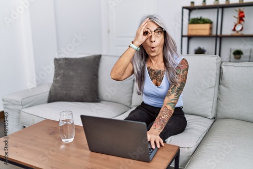 Middle age grey-haired woman using laptop at home doing ok gesture shocked with surprised face, eye looking through fingers. unbelieving expression.