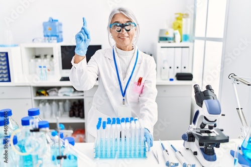 Middle age grey-haired woman wearing scientist uniform smiling with an idea or question pointing finger with happy face, number one
