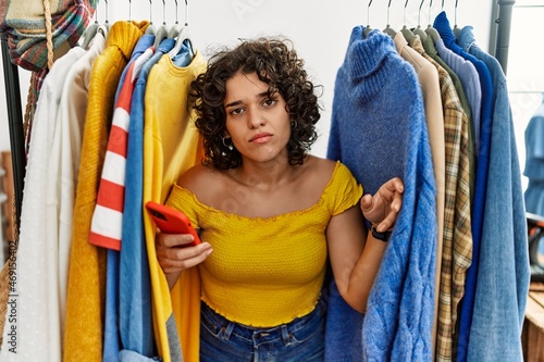 Young hispanic woman searching clothes on clothing rack using smartphone looking sleepy and tired, exhausted for fatigue and hangover, lazy eyes in the morning.