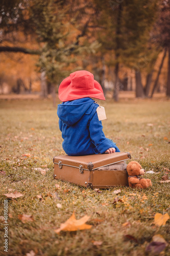 A child in a blue coat and a red hat is sitting on a suitcase. A children's photo shoot in the image of Paddington bear