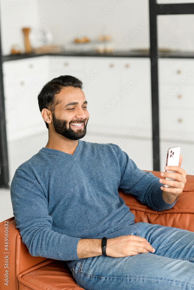 Fototapeta premium Video call concept. Hilarious dark-haired bearded Indian man in casual wear using smartphone for virtual meeting, enjoys online chatting sitting on the sofa at home, guy watching movie and laughing