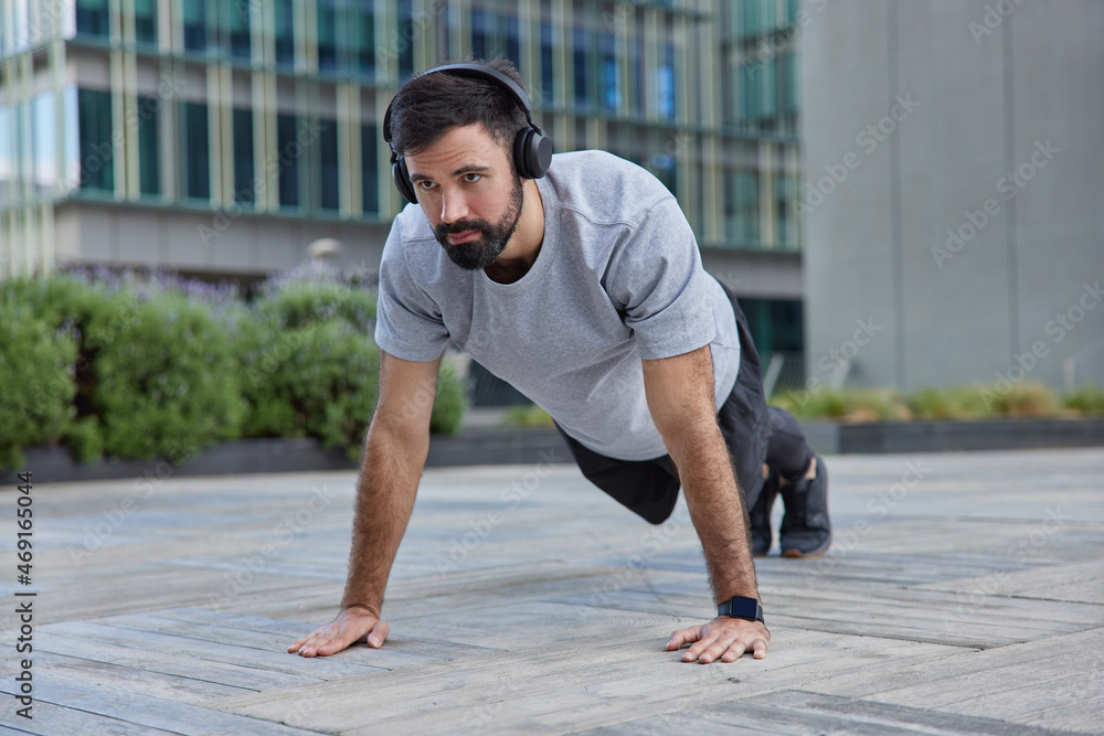 Fototapeta premium Outdoor shot of motivated strong bearded man does plank exercise demonstrates endurance poses against urban background listens music in headphones makes strength workout trains body does push ups