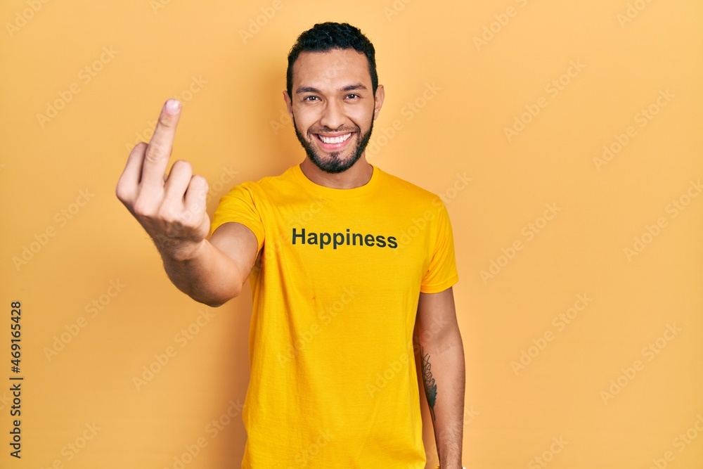 Hispanic man with beard wearing t shirt with happiness word message showing middle finger, impolite and rude fuck off expression Hispanic man with beard wearing t shirt with happiness word message showing middle finger, impolite and rude fuck off expression