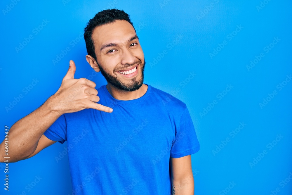 Hispanic man with beard wearing casual blue t shirt smiling doing phone gesture with hand and fingers like talking on the telephone. communicating concepts. Hispanic man with beard wearing casual blue t shirt smiling doing phone gesture with hand and fingers like talking on the telephone. communicating concepts.