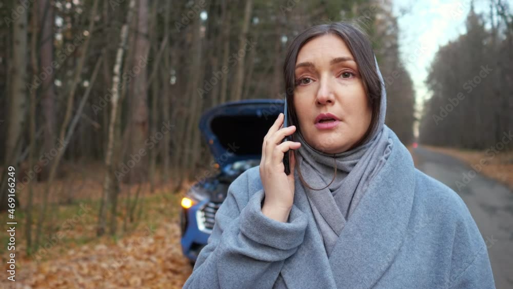 Lady driver in warm coat stands near broken automobile with open hood and calls emergency repair service for help on roadside near autumn forest, closep Lady driver in warm coat stands near broken automobile with open hood and calls emergency repair service for help on roadside near autumn forest, closep
