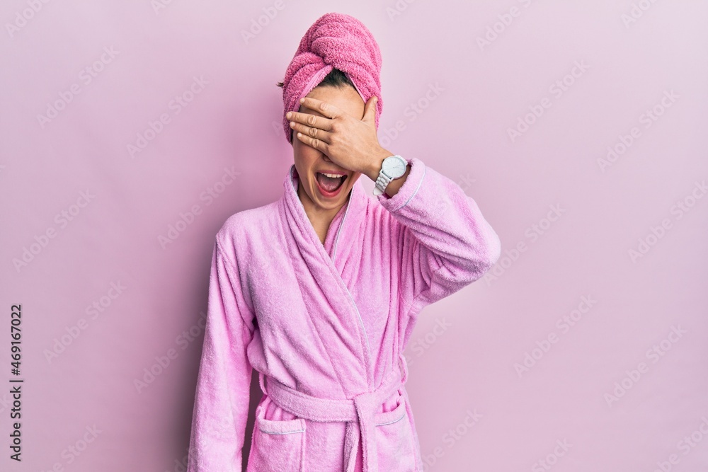 Young blonde woman wearing shower towel cap and bathrobe smiling and laughing with hand on face covering eyes for surprise. blind concept. - obrazy, fototapety, plakaty 