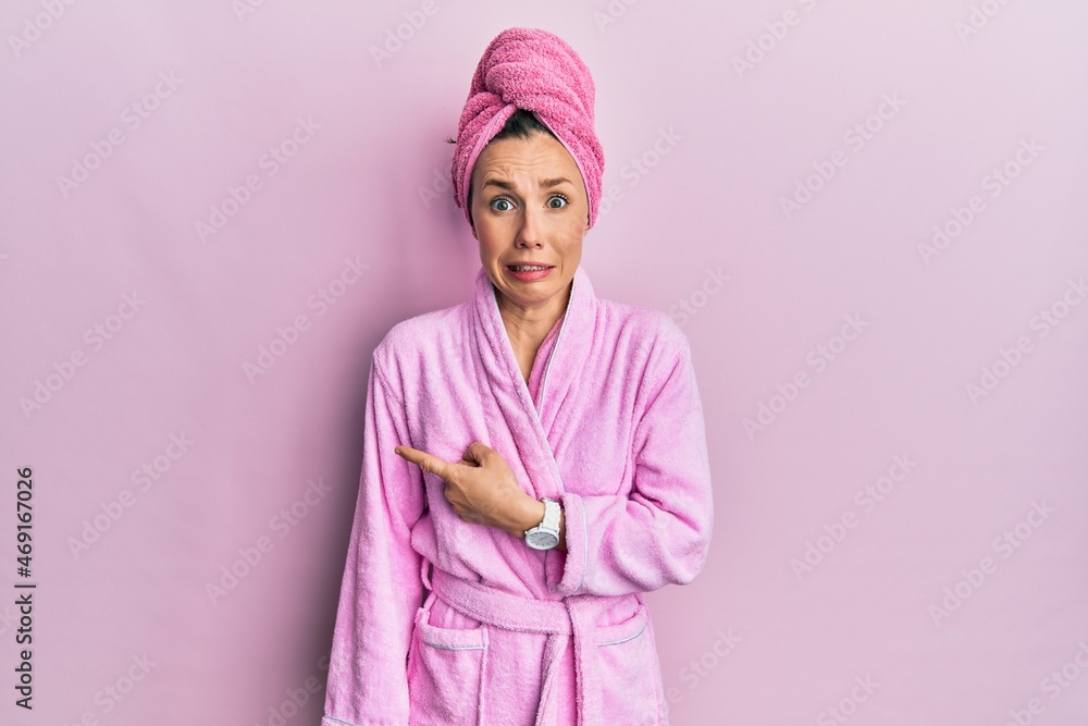 Fototapeta premium Young blonde woman wearing shower towel cap and bathrobe pointing aside worried and nervous with forefinger, concerned and surprised expression