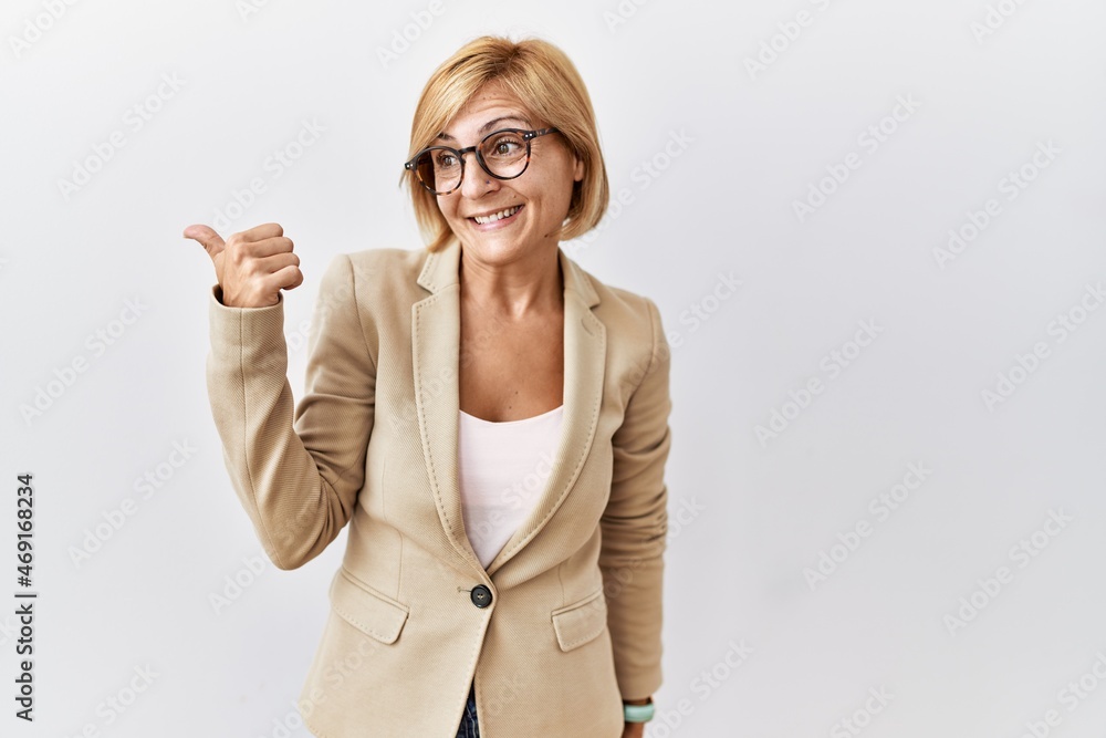 Middle age blonde business woman standing over isolated background smiling with happy face looking and pointing to the side with thumb up. Middle age blonde business woman standing over isolated background smiling with happy face looking and pointing to the side with thumb up.